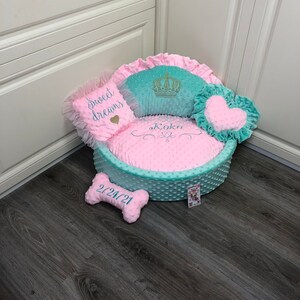 May include: A light blue and pink dog bed with a crown, heart, and bone shaped pillows. The bed has a pink polka dot pattern and the pillows have white embroidery that says "Sweet Dreams", "Koko", and "2/24/21".