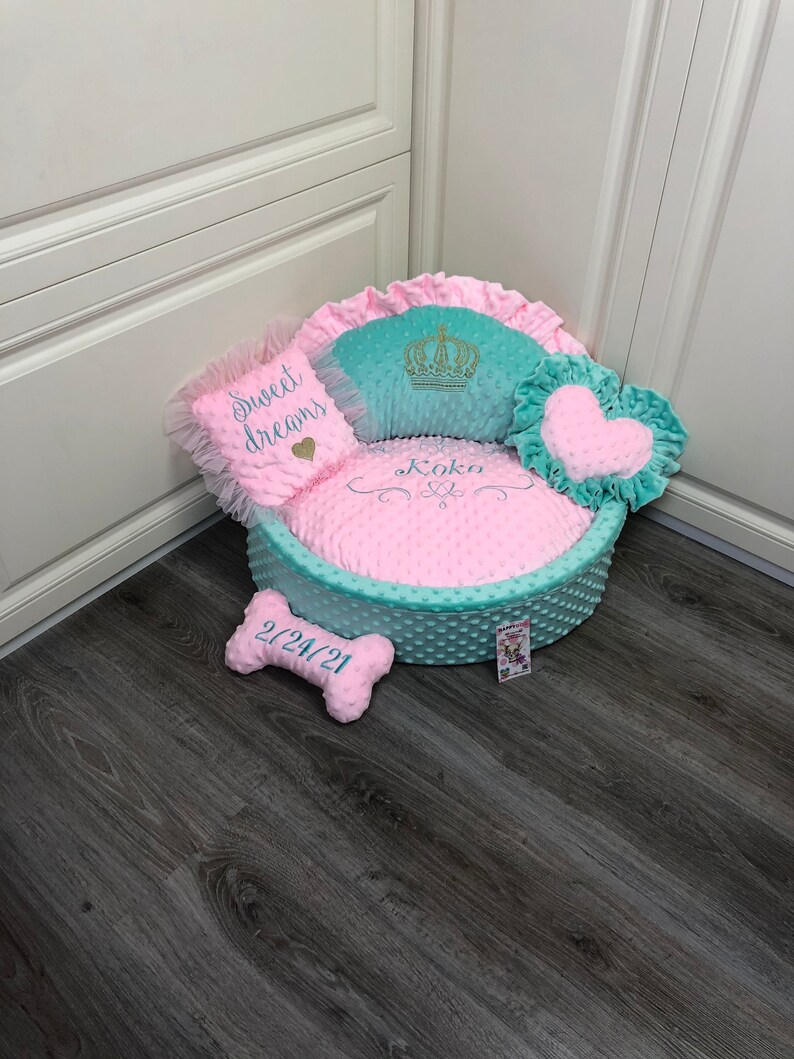 May include: A light blue and pink dog bed with a crown, heart, and bone shaped pillows. The bed has a pink polka dot pattern and the pillows have white embroidery that says "Sweet Dreams", "Koko", and "2/24/21".
