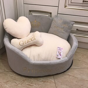May include: A gray and white dog bed with a crown, bone, and heart-shaped pillows. The bed is embroidered with the words "Gucci" and "Princess Gucci".