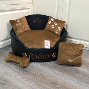 May include: A black and brown pet bed with embroidered text and paw prints. The bed includes two square pillows and a bone-shaped toy. The bed is personalized with the name "Radar".