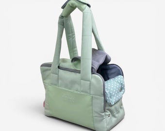 May include: A sage green pet carrier bag with long padded handles, a zippered top opening, and a front pocket. The interior features a light blue lining with white polka dots and a gray mesh panel. The exterior has a smooth, quilted texture.