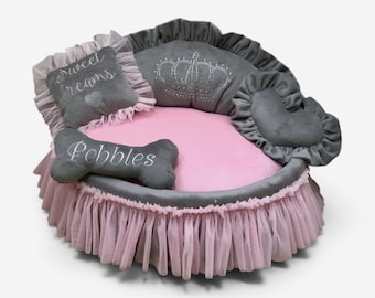 May include: A round pet bed with a pink plush interior and a grey ruffled exterior. It includes a square pillow with the text "sweet dreams", a bone-shaped pillow with the text "Pebbles", and a heart-shaped pillow, all featuring grey fabric.