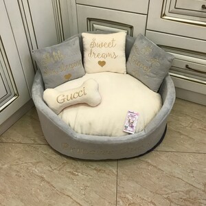 May include: A gray and white dog bed with a plush, cream-colored cushion. The bed has embroidered pillows with the text "Shh... I'm sleeping", "Sweet dreams", "Gucci", and "Gucci". The bed also has a bone-shaped pillow with the text "Gucci" and the words "Princess Gucci" embroidered on the side.