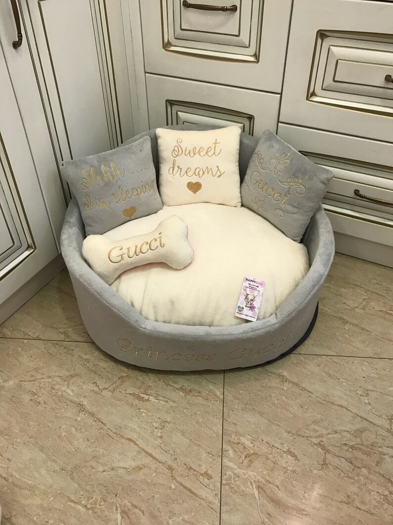 May include: A gray and white dog bed with a plush, cream-colored cushion. The bed has embroidered pillows with the text "Shh... I'm sleeping", "Sweet dreams", "Gucci", and "Gucci". The bed also has a bone-shaped pillow with the text "Gucci" and the words "Princess Gucci" embroidered on the side.