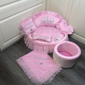 May include: Pink princess-themed dog bed with a crown, a heart-shaped pillow, a bone-shaped pillow, and a blanket. The bed has a ruffled trim and the blanket has a ruffled edge. The bed is on a wooden floor.
