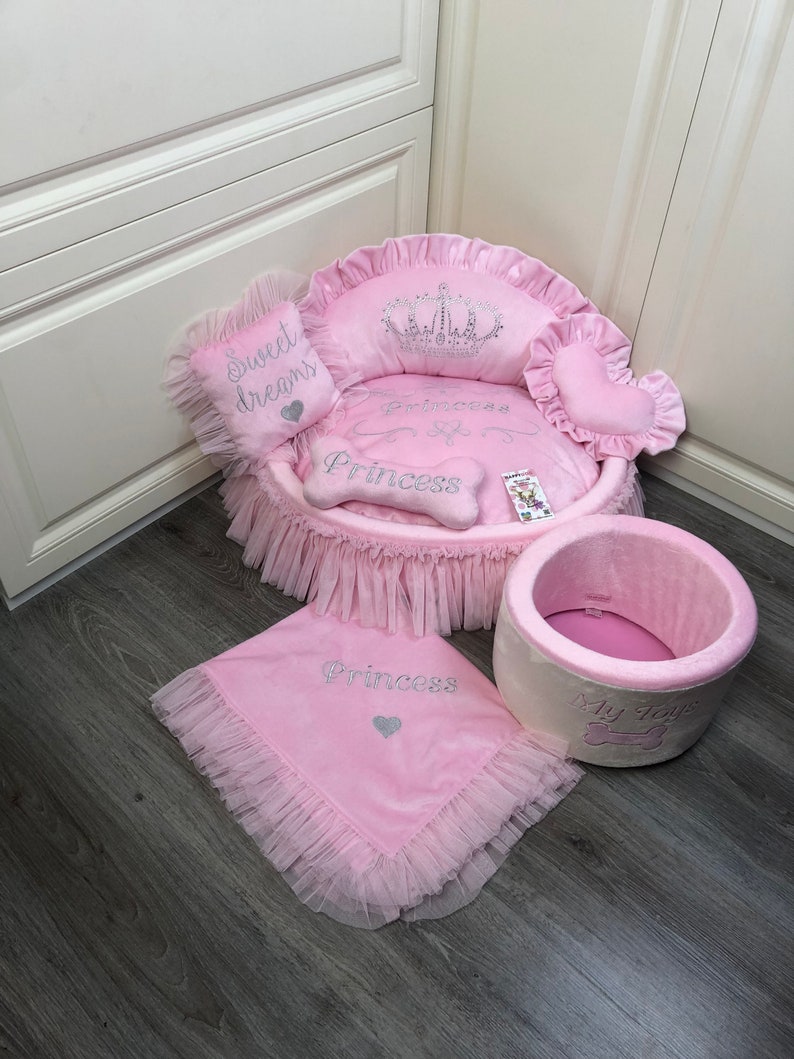 May include: Pink princess-themed dog bed with a crown, a heart-shaped pillow, a bone-shaped pillow, and a blanket. The bed has a ruffled trim and the blanket has a ruffled edge. The bed is on a wooden floor.
