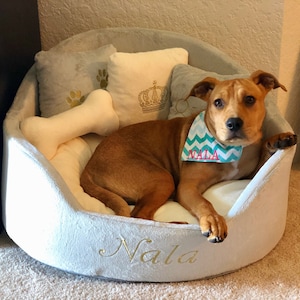 May include: A brown and white dog with a turquoise bandana that says "Nala" is lying in a light gray dog bed with a bone-shaped pillow. The bed is embroidered with the name "Nala".