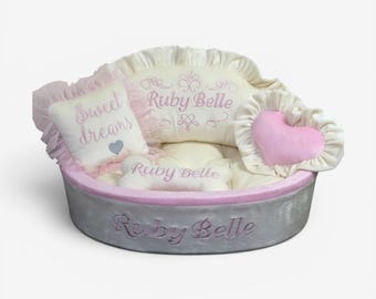 May include: A round gray pet bed with a pink trim, featuring a cream-colored back cushion embroidered with "Ruby Belle," a square pillow with "Sweet dreams" and a heart, a pink heart-shaped pillow, and a bone-shaped toy labeled "Ruby Belle."
