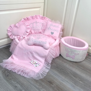 May include: Pink dog bed with a crown pillow, a heart-shaped pillow that says "Sweet Dreams", and a smaller pillow that says "Princess Milana". The bed has a pink and white tulle skirt. There is also a pink and white dog toy bin that says "My Toys" with a bone graphic.