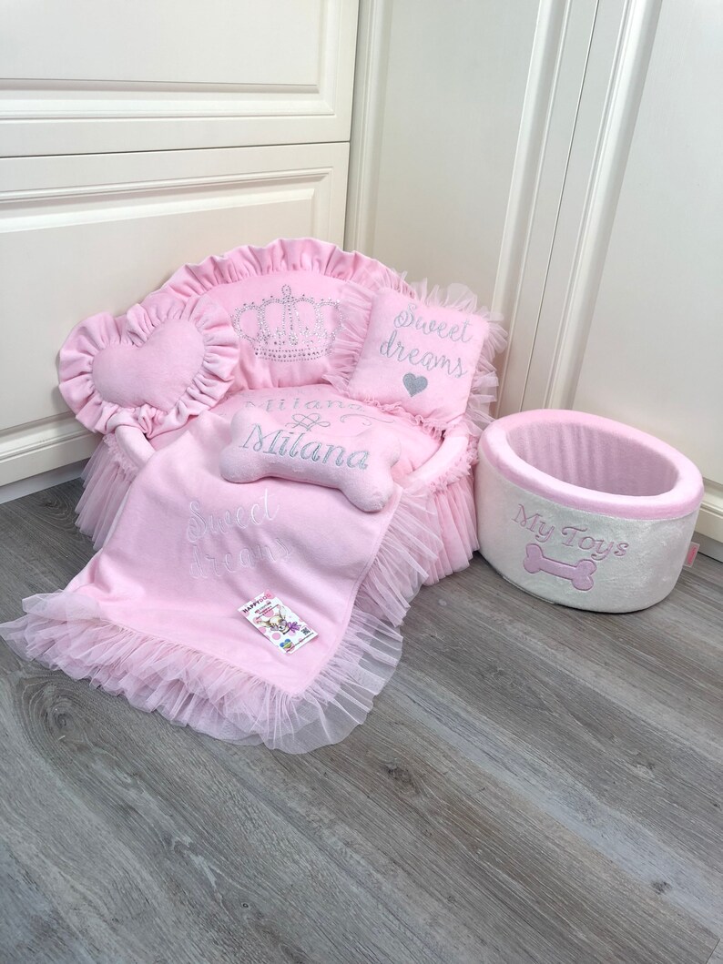 May include: Pink dog bed with a crown pillow, a heart-shaped pillow that says "Sweet Dreams", and a smaller pillow that says "Princess Milana". The bed has a pink and white tulle skirt. There is also a pink and white dog toy bin that says "My Toys" with a bone graphic.