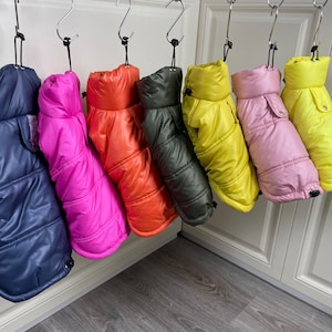 May include: Seven dog puffer jackets hanging on hangers. The jackets are in various colours, including navy blue, pink, orange, green, yellow, and pink.
