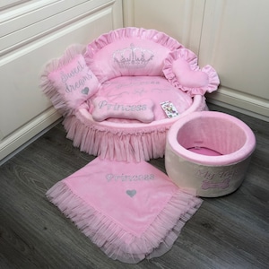 May include: Pink dog bed with a crown and the word "Princess" embroidered on it. The bed has a ruffled edge and comes with a matching blanket and a pink and white toy bin with the words "My Toys" on it.