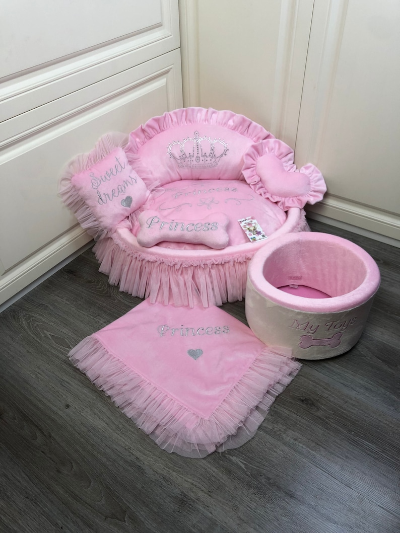 May include: Pink dog bed with a crown and the word "Princess" embroidered on it. The bed has a ruffled edge and comes with a matching blanket and a pink and white toy bin with the words "My Toys" on it.