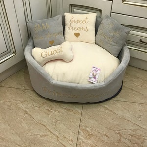 May include: A gray and white dog bed with a bone-shaped pillow that says "Gucci" in gold lettering. The bed has three pillows with the text "Shh... I'm sleeping", "Sweet dreams" and "Gucci" in gold lettering. The bed also has the text "Princess Gucci" in gold lettering.