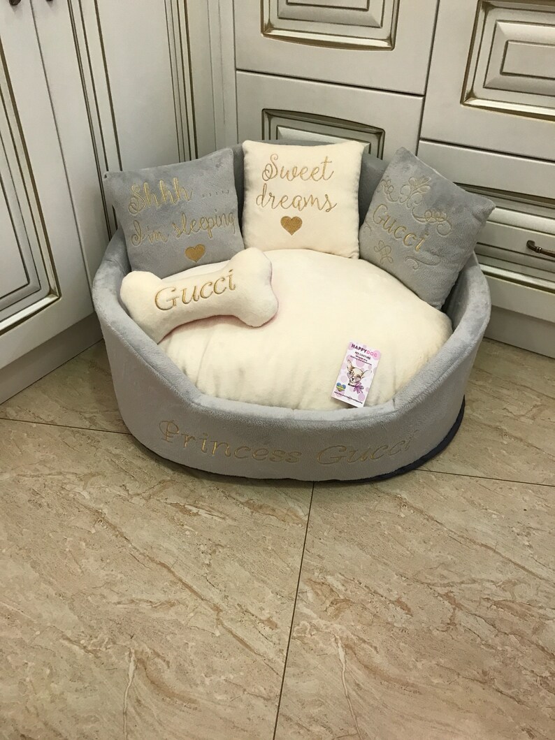May include: A gray and white dog bed with a bone-shaped pillow that says "Gucci" in gold lettering. The bed has three pillows with the text "Shh... I'm sleeping", "Sweet dreams" and "Gucci" in gold lettering. The bed also has the text "Princess Gucci" in gold lettering.
