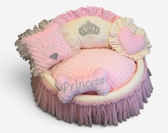 May include: A round, pink pet bed with a ruffled skirt, featuring a dimpled minky fabric texture. It includes a square pillow with the text "Princess's Dog" and a heart, a bone-shaped pillow with "Princess" embroidered, and a crown motif.