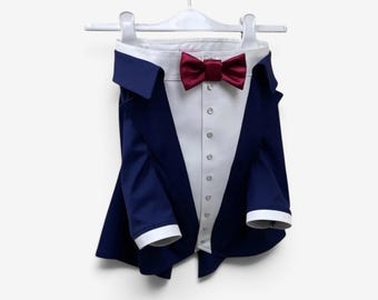 Puede incluir: Una chaqueta estilo esmoquin azul marino con un frente de camisa blanco, una fila vertical de botones pequeños y una pajarita de satén rojo oscuro. La chaqueta tiene cuello y puños blancos en las mangas largas, colgada en una percha blanca.