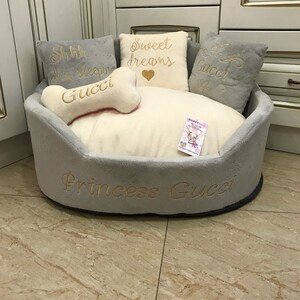 May include: A gray and white dog bed with a plush, white interior. The bed has a gray border with the text "Princess Gucci" embroidered in gold. There are four pillows inside the bed, two gray and two white. The white pillows have gold embroidery that reads "Sweet dreams" and "Shhh... I'm sleeping Gucci".