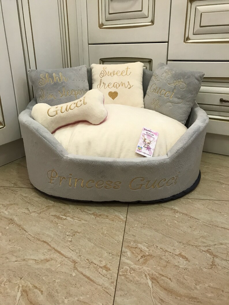 May include: A gray and white dog bed with a plush, white interior. The bed has a gray border with the text "Princess Gucci" embroidered in gold. There are four pillows inside the bed, two gray and two white. The white pillows have gold embroidery that reads "Sweet dreams" and "Shhh... I'm sleeping Gucci".