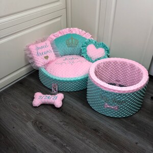 May include: A pink and teal dog bed with a heart-shaped pillow, a crown pillow, and a bone-shaped pillow. The bed has the words "Koko" and "My Toys" embroidered on it.