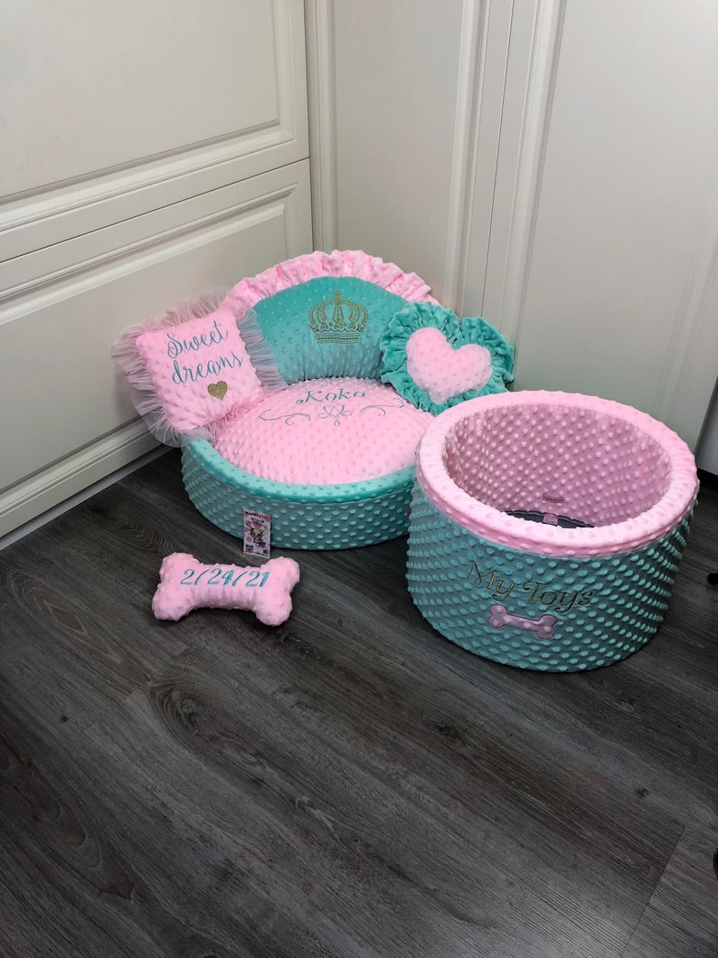 May include: A pink and teal dog bed with a heart-shaped pillow, a crown pillow, and a bone-shaped pillow. The bed has the words "Koko" and "My Toys" embroidered on it.