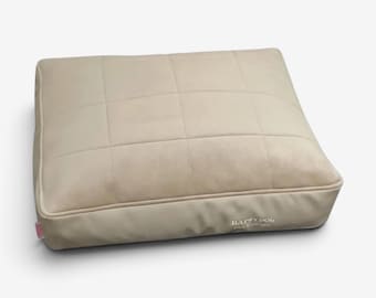 Puede incluir: Una cama rectangular para mascotas de color beige con una superficie superior acolchada en un patrón de cuadrícula. El lateral de la cama incluye una pequeña etiqueta de tela rosa y el texto bordado en blanco Happy Dog Pet Couture.