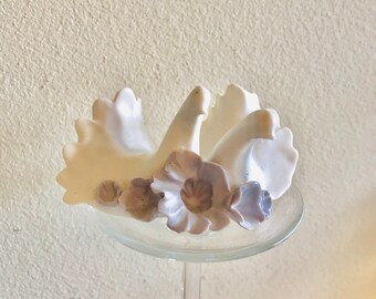 May include: A white ceramic sculpture featuring two stylized birds with fanned tail feathers, adorned with a cluster of pale lavender and light gray sculpted flowers at the base, all resting on a clear glass pedestal stand with a circular top.