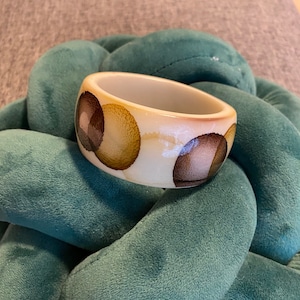 May include: A wide bangle bracelet with a smooth, cream-colored base featuring a repeating pattern of overlapping circular shapes in shades of brown, tan, and dark chocolate. The bracelet has a glossy finish and a thick, rounded profile.