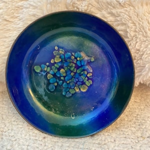 May include: A small, round, enameled dish with a vibrant color palette. The dish features a deep blue center that transitions to a green rim. The center has a cluster of blue and yellow shapes. The dish has a metallic rim.