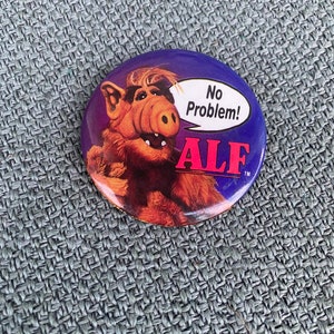 May include: A round button featuring the character ALF from the 1980s sitcom. The button has a purple background with ALF's image and the words "No Problem!" in a speech bubble. The word "ALF" is printed in red.