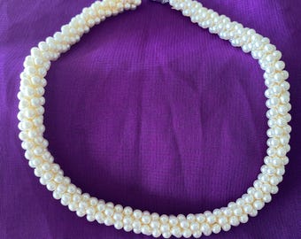 May include: Cream-colored pearl necklace with a silver clasp, resting on a purple fabric. The necklace is made of small, round pearls, tightly strung together to form a thick, elegant strand. A classic jewelry piece.