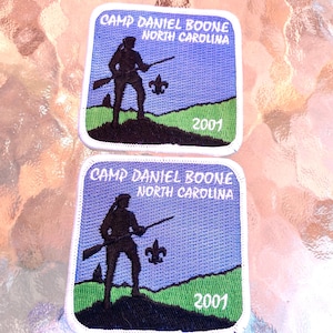 May include: Two embroidered patches with a blue background and the text "CAMP DANIEL BOONE NORTH CAROLINA" at the top. A silhouette of a person holding a rifle is in the center, with a fleur-de-lis below. The year "2001" is at the bottom.