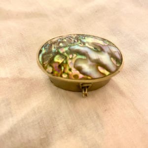 May include: An oval-shaped trinket box with a shimmering abalone shell top. The box has a gold-colored metal base and a small clasp. The abalone shell displays iridescent colors of green, blue, and purple.