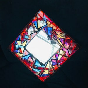 May include: A square mirror with a colorful mosaic frame. The frame is made of triangular pieces in shades of red, blue, gold, and purple. The mirror is set against a black background.