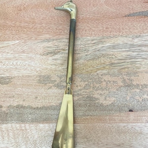 May include: A long-handled shoehorn made of polished brass. The handle features a detailed duck head at the top, transitioning into a slender cylindrical shaft that connects to a wide, curved scoop at the bottom for sliding into footwear.