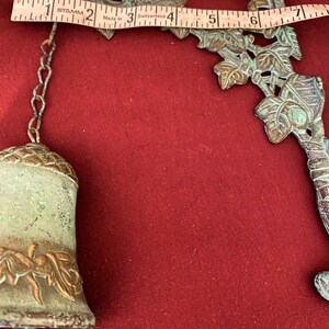 Pode incluir: Um sino de metal verde com um design de folhas pendura em uma corrente presa a um suporte de parede. O suporte de parede tamb&eacute;m &eacute; feito de metal verde e apresenta um design de folhas.