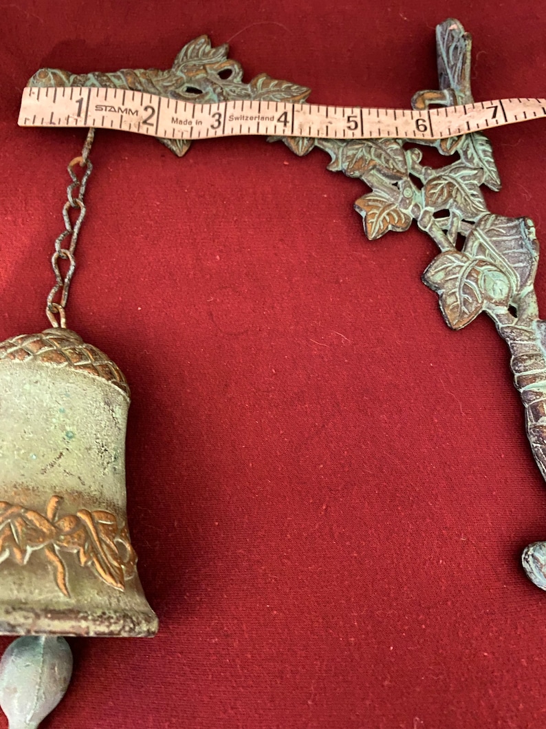 Pode incluir: Um sino de metal verde com um design de folhas pendura em uma corrente presa a um suporte de parede. O suporte de parede tamb&eacute;m &eacute; feito de metal verde e apresenta um design de folhas.