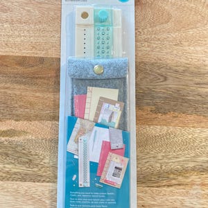 May include: A book binding guide kit in a clear plastic package. The kit includes a gray felt pouch with a gold snap, a teal and white tool, and various paper inserts. The packaging has text in multiple languages.