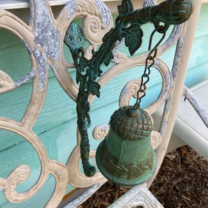 Pode incluir: Um sino de metal verde com um design de folhas pendura em um suporte decorativo de metal. O suporte est&aacute; preso a uma cerca de metal branco com um padr&atilde;o floral.