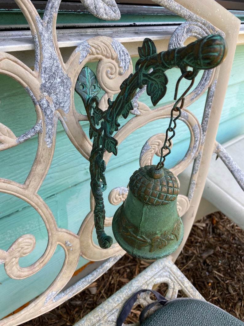 Pode incluir: Um sino de metal verde com um design de folhas pendura em um suporte decorativo de metal. O suporte est&aacute; preso a uma cerca de metal branco com um padr&atilde;o floral.