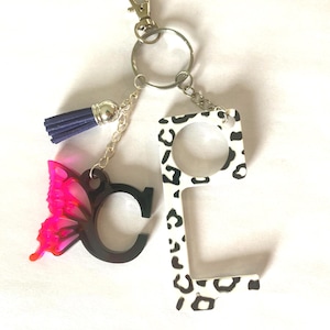 May include: A keychain featuring a silver-tone metal ring and lobster clasp, a dark blue tassel, a black letter C charm attached to a translucent pink butterfly wing, and a white L-shaped tool with a black leopard print pattern throughout.