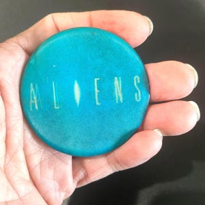 May include: A round, teal button with the word "ALIENS" printed in white. The button has a textured surface and a vertical white line between the letters. The button is a collectible item.