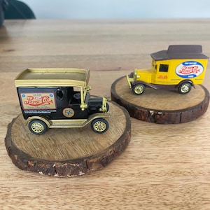 May include: Two miniature vintage Pepsi Cola delivery trucks, one black and one yellow, sit on wooden coasters. The black truck has gold accents and the text "Drink Pepsi Cola" and "5¢". The yellow truck has the text "You Can Get Pepsi Here".