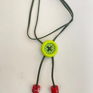 May include: A unique bolo tie featuring a lime green button-shaped pendant with a central design of green rhinestones. The tie has a dark green cord and red cylindrical end pieces. This handcrafted accessory adds a pop of color.