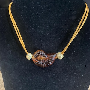 May include: A necklace featuring a dark amber-colored glass pendant shaped like an ammonite fossil. The pendant is flanked by two small, round, cream and blue striped glass beads, all suspended on a double-stranded mustard yellow cord.