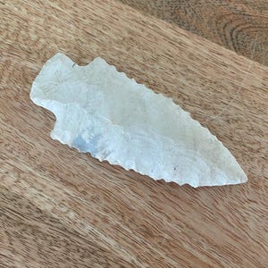 May include: A translucent, off-white stone arrowhead featuring a sharp, pointed tip and serrated edges along its length. The object has a distinct notched base and a flaked, textured surface characteristic of a hand-knapped stone projectile point.