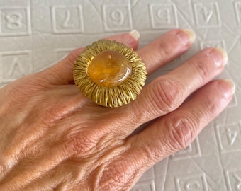 May include: A gold-toned ring with a large, oval, amber-colored stone. The ring has a textured, sunburst-like design around the stone. The ring is on a person's hand.