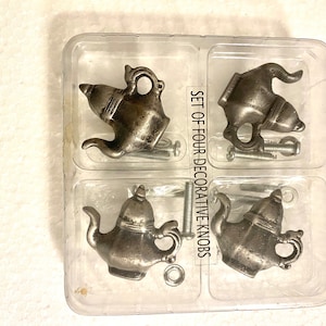 May include: Set of four decorative teapot-shaped cabinet knobs in a clear plastic packaging. The knobs are silver in color and have a vintage design. The packaging has the text "Set of Four Decorative Knobs" printed on it.