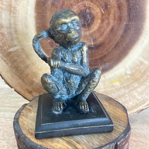 May include: A bronze-toned monkey figurine in a seated position with its tail curved upward. The monkey is mounted on a square black base, featuring detailed fur texture and facial features, capturing a realistic posture with one hand near its head.
