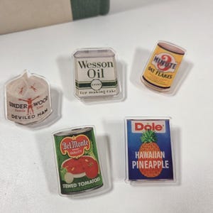 May include: A collection of five clear acrylic pins featuring vintage food product designs. The pins include images of Underwood Deviled Ham, Wesson Oil, 3-Minute Oat Flakes, Del Monte Stewed Tomatoes, and Dole Hawaiian Pineapple.
